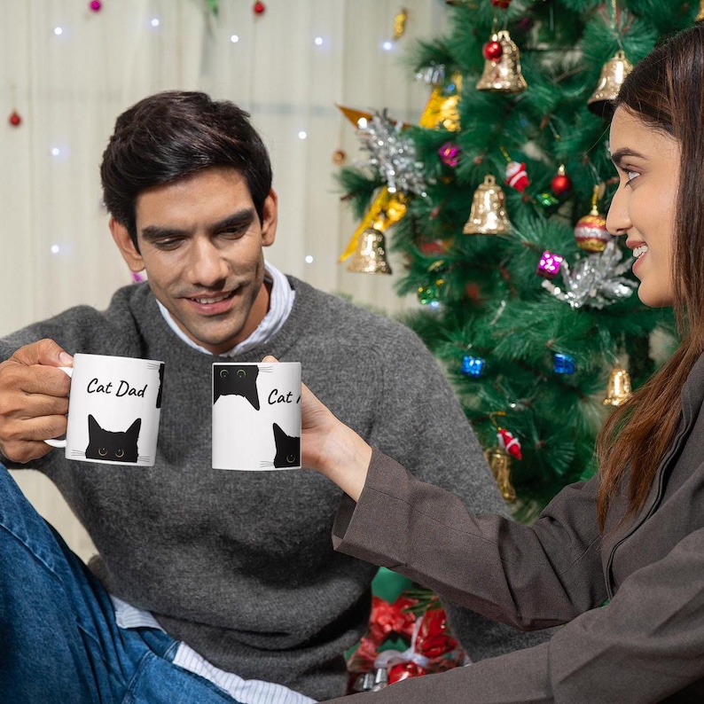 May include: Two white ceramic mugs featuring black cat designs. One mug says "Cat Dad" and the other says "Cat". The mugs are held by two people, with a decorated Christmas tree in the background.