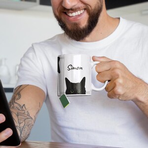 May include: White ceramic mug with a black cat design and the name "Simon" in black script. The cat's eyes are visible. A green tea bag is attached. The mug is held by a person.
