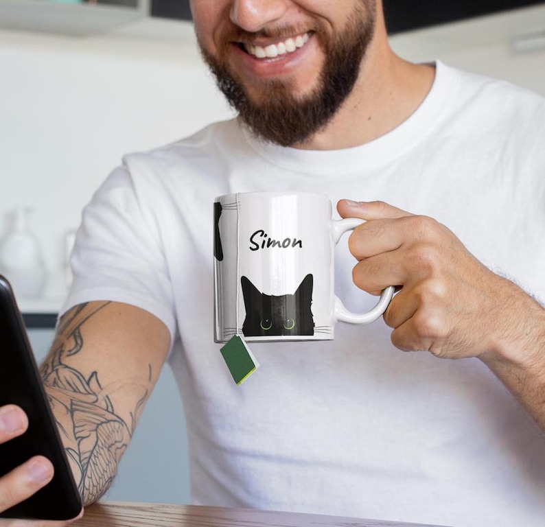 May include: White ceramic mug with a black cat design and the name "Simon" in black script. The cat's eyes are visible. A green tea bag is attached. The mug is held by a person.