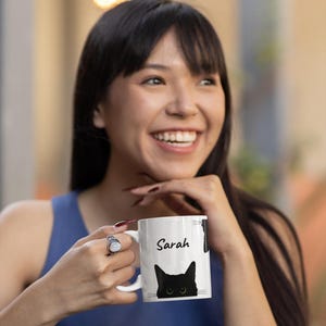 May include: White ceramic mug with a black cat design and the name "Sarah" in black script. The cat has green eyes. The mug has a curved handle and a glossy finish. The cat's face is visible at the bottom of the mug.