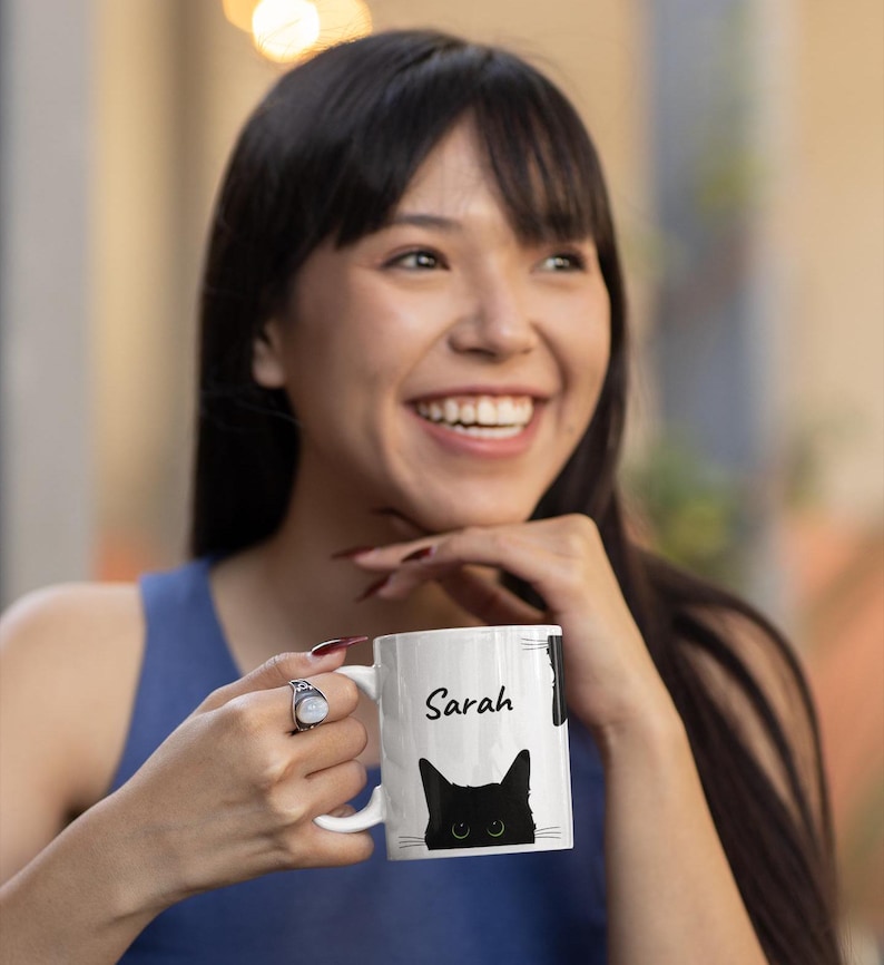 May include: White ceramic mug with a black cat design and the name "Sarah" in black script. The cat has green eyes. The mug has a curved handle and a glossy finish. The cat's face is visible at the bottom of the mug.
