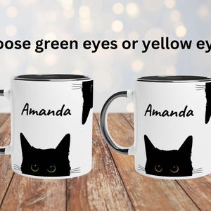 May include: Two white ceramic mugs with black rims and handles, each with a black cat design. One mug has green eyes, the other yellow. The name "Amanda" is printed on each mug. Text above reads "Choose green eyes or yellow eyes."