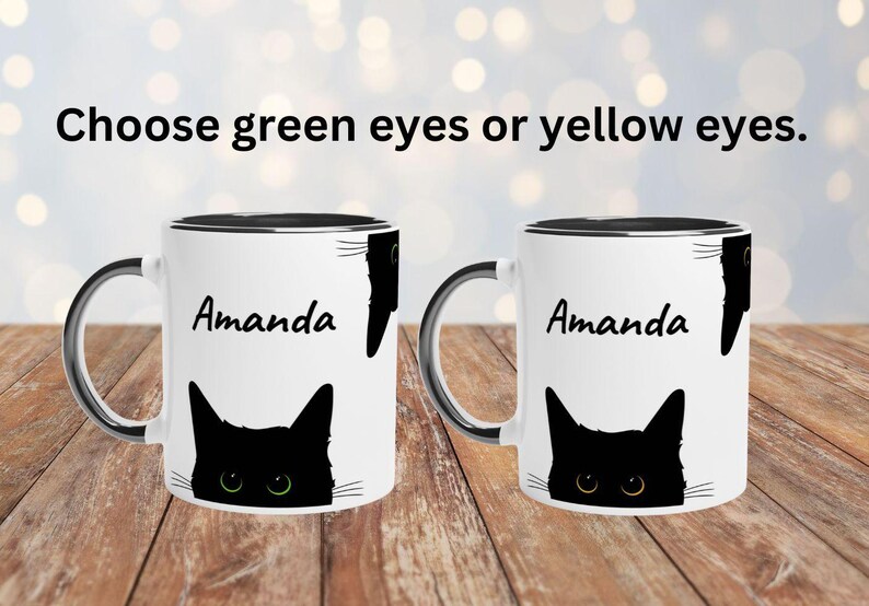 May include: Two white ceramic mugs with black rims and handles, each with a black cat design. One mug has green eyes, the other yellow. The name "Amanda" is printed on each mug. Text above reads "Choose green eyes or yellow eyes."