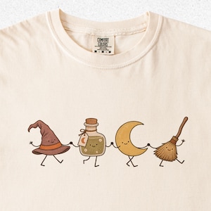 May include: A cream-colored t-shirt with a Comfort Colors label at the neck. The graphic features a brown witch hat, a potion bottle, a yellow crescent moon, and a broomstick, all with smiling faces and stick limbs, holding hands in a line.
