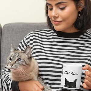 May include: White ceramic mug with the text "Cat Mom" and a black cat silhouette with green eyes. The mug is held by a person wearing a black and white striped top, with a grey cat in their arms.