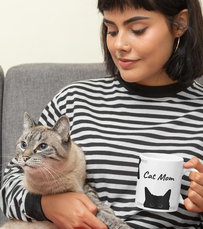 May include: White ceramic mug with the text "Cat Mom" and a black cat silhouette with green eyes. The mug is held by a person wearing a black and white striped top, with a grey cat in their arms.