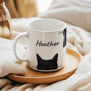 May include: White ceramic mug featuring a black cat face design and the name "Heather" printed in black. The mug sits on a wooden tray, with a blurred background.