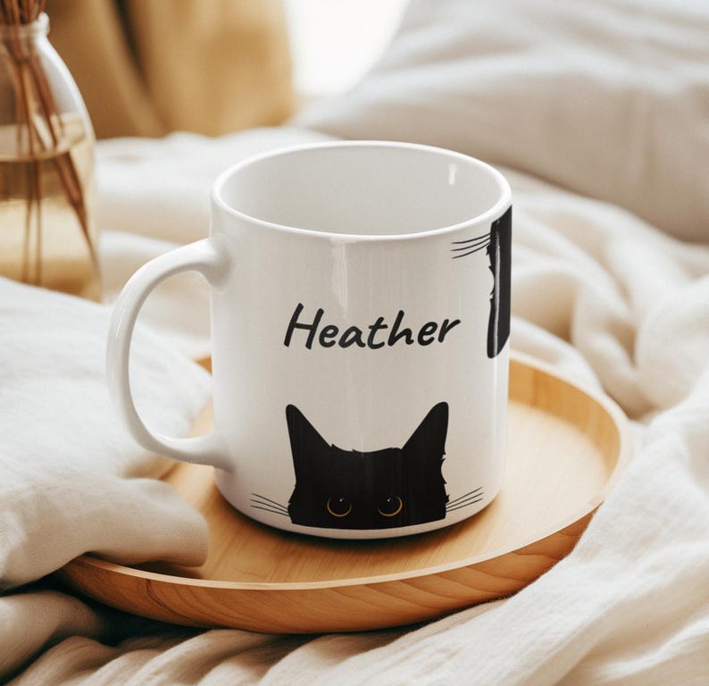 May include: White ceramic mug featuring a black cat face design and the name "Heather" printed in black. The mug sits on a wooden tray, with a blurred background.