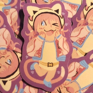 May include: A stack of stickers featuring a cartoon character with pink hair, wearing a white jacket and a yellow cat-eared hat. The character is winking and has a playful expression. The stickers are on a purple background with white stars.