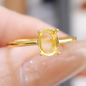 May include: A gold-colored ring with an oval-shaped setting. The ring is held by a person. The setting has four prongs to hold a gemstone. The ring's band is thin and smooth.