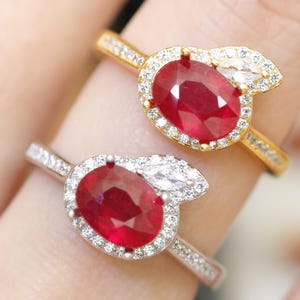 Puede incluir: Dos anillos con piedras preciosas ovaladas de color rojo intenso. Un anillo es dorado con un halo de pequeños diamantes y un acento de diamante blanco talla marquesa. El otro anillo es plateado con un diseño similar.