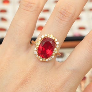 Puede incluir: Un anillo de oro con una gran piedra preciosa de color rojo intenso y corte ovalado en el centro, rodeada por un halo de pequeñas piedras blancas claras y redondas engastadas en un marco de oro festoneado. El aro es fino y de oro pulido.