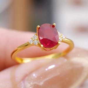 Op de afbeelding: Een gouden ring met een grote, ovaal geslepen robijn. De robijn heeft een dieprode kleur en wordt aan weerszijden geflankeerd door kleinere, heldere edelstenen. De ring heeft een delicate, gepolijste gouden band.