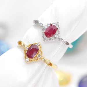 Könnte beinhalten: Zwei Ringe mit ovalen, roten Edelsteinen. Ein Ring ist goldfarben mit dekorativer Fassung und kleinen Diamanten. Der andere Ring ist silberfarben mit ähnlicher Fassung und kleinen Diamanten. Beide Ringe sind auf weißem Hintergrund.