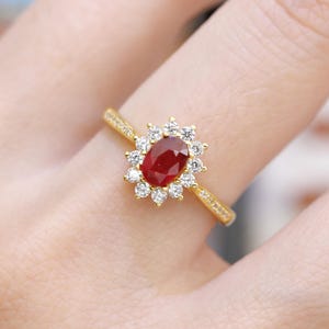 May include: A gold ring featuring a large oval-cut ruby gemstone surrounded by a halo of small, round, clear gemstones. The ring's band is also adorned with small gemstones, creating a sparkling effect. The ring is displayed on a finger.
