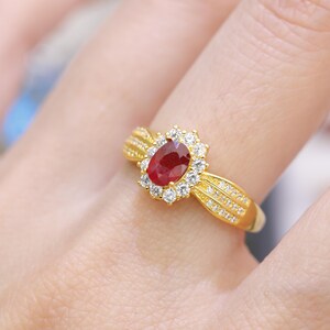 Pode incluir: Um anel de ouro com uma grande pedra de rubi ovalada rodeada por pequenas pedras redondas e transparentes. A banda do anel tem um design texturizado com pequenas pedras adicionais. O anel é exibido num dedo.