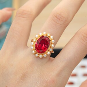 Op de afbeelding: Een gouden ring met een grote ovaal geslepen rode edelsteen als middelpunt, omringd door een halo van kleine witte parels en piepkleine doorzichtige steentjes. De metalen ring heeft een textuurrijke, decoratieve afwerking rondom.