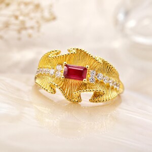 May include: A gold ring featuring a central rectangular baguette-cut red gemstone. The band has a textured, fan-like design with radiating lines and is accented by small, clear round stones set along the sides of the central red gemstone.
