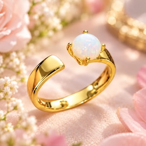 Può includere: Un anello aperto color oro con un cabochon rotondo in opale bianco iridescente montato su quattro griffe. La fascia presenta una finitura liscia e lucida con un design a apertura regolabile, che mette in risalto la superficie e la gemma luminosa.