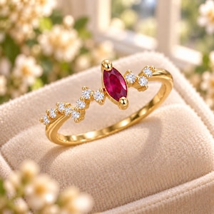 Può includere: Un anello in oro con una pietra preziosa rossa centrale a taglio marquise, affiancata da una serie di piccole pietre bianche rotonde e trasparenti disposte in un delicato motivo asimmetrico lungo la sottile fascia in oro lucido.