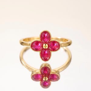 May include: A gold ring featuring a four-petaled flower motif composed of four oval-cut vibrant pink rubies surrounding a central gold bead. The setting has a delicate beaded edge, and the band is a smooth, polished gold metal finish.