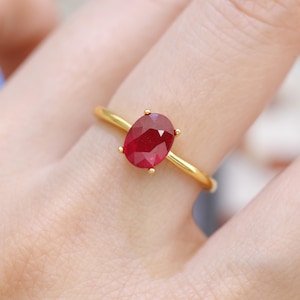 May include: A gold ring featuring a large, oval-cut ruby gemstone. The ruby is a deep red color and is set with four small prongs. The ring's band is a simple, polished gold.