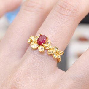 Op de afbeelding: Een goudkleurige ring met een ovale robijn in het midden. De ring is versierd met bloemmotieven, met kleine, heldere edelstenen op de bloemblaadjes. De band heeft een delicaat, door de natuur geïnspireerd ontwerp.