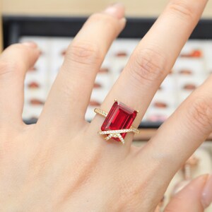 May include: A gold ring featuring a large, rectangular, emerald-cut red gemstone as the centerpiece. The band is composed of two thin, crossing gold wires encrusted with small, clear, sparkling stones that wrap around the base of the red stone.