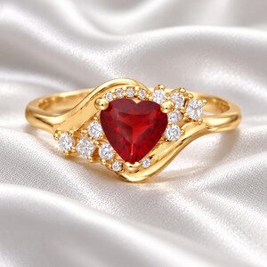 May include: A gold ring featuring a central heart-shaped red gemstone surrounded by a cluster of small, clear, round and square-cut white stones. The band has a smooth, polished gold finish that curves around the central stone setting.