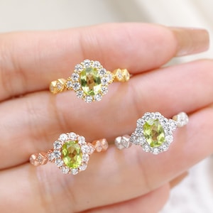May include: Three rings with oval, light green gemstones. Each ring has a halo of small, clear stones around the center stone. The bands are gold, rose gold, and silver. The rings are displayed on a light background.