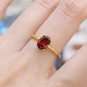 May include: A gold ring with a striking, oval-shaped, deep red gemstone. The gemstone is held in place by four prongs, and the band is plain, polished gold. The ring is displayed on a person's finger.