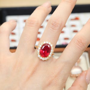 Puede incluir: Un anillo de oro con una gran piedra preciosa de color rojo vibrante y corte ovalado en el centro, rodeada por un halo de pequeñas piedras blancas redondas y transparentes. La banda es fina y de metal dorado, contrastando con la piedra roja.
