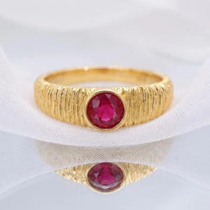 Puede incluir: Un anillo de oro con una piedra preciosa roja vibrante, redonda y facetada, engastada en el centro. La banda presenta una textura vertical similar a la corteza que recorre toda la circunferencia, creando un contraste con el engaste pulido.