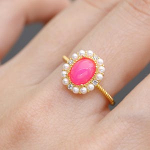 May include: A gold ring featuring a vibrant pink oval gemstone, surrounded by small pearls and sparkling accents. The ring's band has a textured design, adding to its elegant appeal. This piece of jewelry is a stylish accessory.
