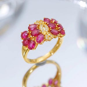 Op de afbeelding: Een gouden ring met een cluster van ovaal geslepen dieproze-rode edelstenen in een bloemachtig patroon, geaccentueerd door kleine heldere witte steentjes in een centraal bladervormig goudmotief, met een gepolijste gouden ringband.