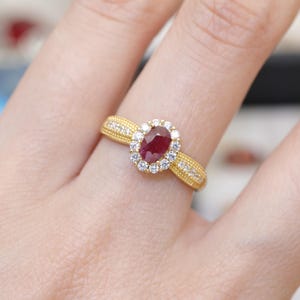 Puede incluir: Un anillo de oro con una piedra central de rubí ovalada, rodeada de pequeños diamantes redondos. La banda tiene un diseño texturizado con diamantes pequeños adicionales. El anillo se muestra en un dedo.