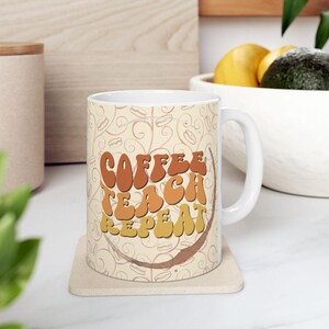 以下が含まれることがあります： 白いセラミック製のコーヒーカップ。茶色、オレンジ、黄色のデザインで、「Coffee Teach Repeat」と書かれています。カップはベージュのコースターの上に置かれています。