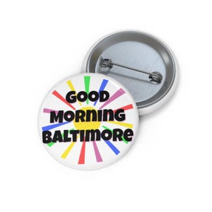 May include: A white button with the text "GOOD MORNING BALTIMORE" in black, with colorful rays emanating from a yellow center. A second button is visible in the background.