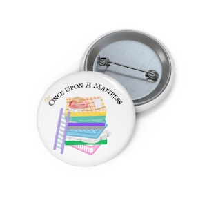 May include: A white button featuring a colourful illustration of a stack of mattresses and a ladder. The text "Once Upon A Mattress" is written in black around the image.