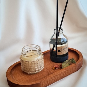 Op de afbeelding: Een houten ovaal dienblad met een kleine glazen potkaars met een geweven jute wikkel, een glazen diffuserfles met zwarte rietjes en een kleine gedroogde bloem. De diffuserfles heeft een label met de tekst "ARTEMIS".