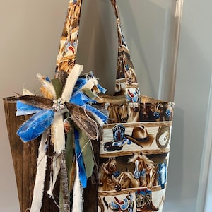 May include: A tote bag featuring a brown wood-grain side and a patterned side with horses, cowboy boots, hats, and lassos. The bag includes two shoulder straps and a decorative cluster of frayed fabric ribbons in blue, white, and olive green.