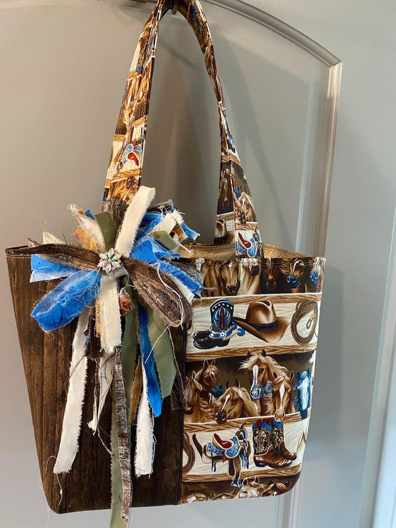 May include: A tote bag featuring a brown wood-grain side and a patterned side with horses, cowboy boots, hats, and lassos. The bag includes two shoulder straps and a decorative cluster of frayed fabric ribbons in blue, white, and olive green.
