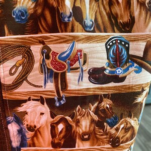 May include: A fabric panel featuring a repeating western pattern with rows of horse heads in various shades of brown, white, and tan, interspersed with illustrations of leather saddles, coiled lassos, and blue-accented cowboy boots.