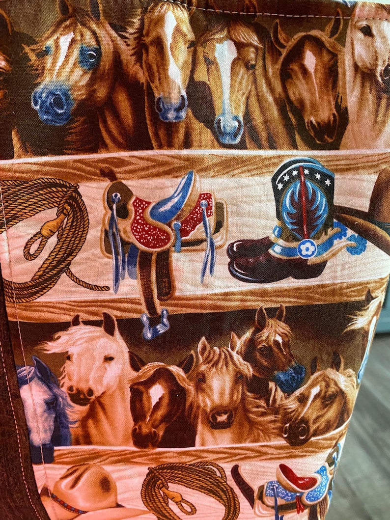 May include: A fabric panel featuring a repeating western pattern with rows of horse heads in various shades of brown, white, and tan, interspersed with illustrations of leather saddles, coiled lassos, and blue-accented cowboy boots.