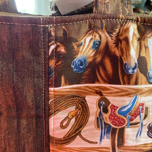 May include: A fabric tote bag featuring a repeating pattern of brown horses, western saddles, and coiled ropes. The bag has a dark brown wood-grain textured side panel and a main section with a light-colored background, all joined by visible stitching.