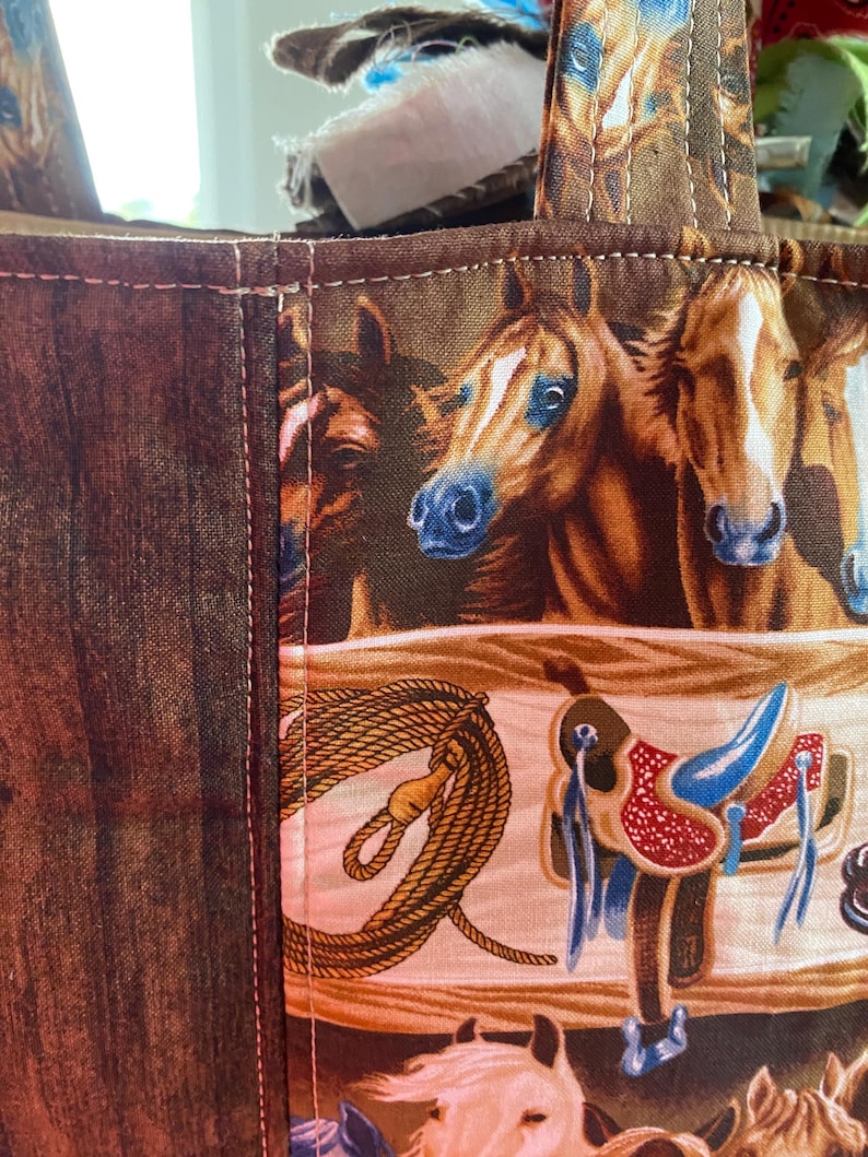 May include: A fabric tote bag featuring a repeating pattern of brown horses, western saddles, and coiled ropes. The bag has a dark brown wood-grain textured side panel and a main section with a light-colored background, all joined by visible stitching.