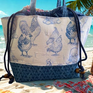 May include: A white and blue tote bag with a chicken print. The bag has a drawstring closure and navy blue rope handles. The bottom of the bag is navy blue with a patterned design. The bag is on a sandy beach with the ocean in the background.