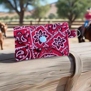 closeup of a red bandana small snap wallet on a piece of rough wood with a ranch and horses in the background.