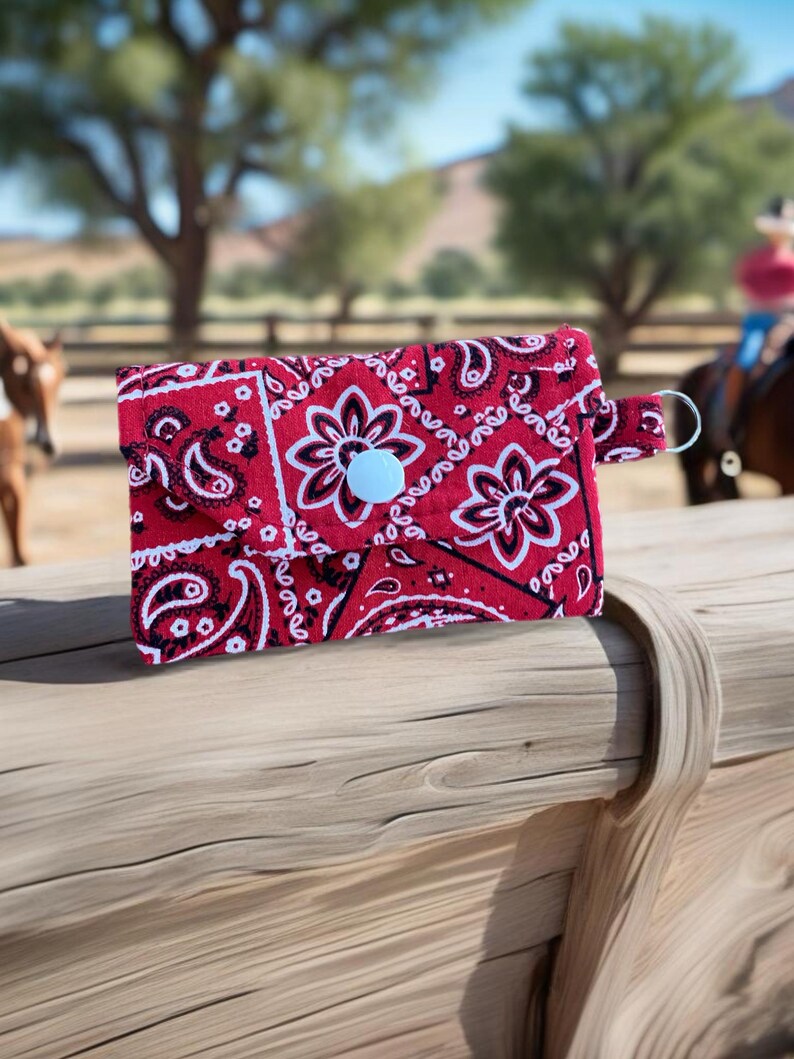 closeup of a red bandana small snap wallet on a piece of rough wood with a ranch and horses in the background.