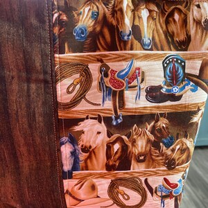 May include: A fabric tote bag featuring a repeating pattern of horses, western saddles, cowboy boots, lassos, and hats. The design includes a dark brown wood-grain textured side panel and a main section with horizontal rows of equestrian imagery.
